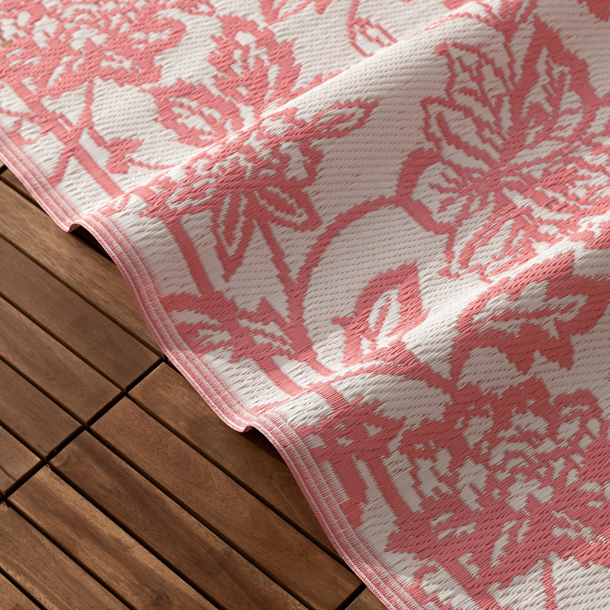 Anemone Jacquard Floral Rug in coral pink and off-white, showing the woven scrolling floral pattern draped over wooden outdoor decking.