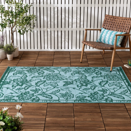 Anemone Jacquard Floral Rug in teal/mineral with Jacobean-style floral pattern and border, shown on a wooden deck beside a woven chair and potted plants.