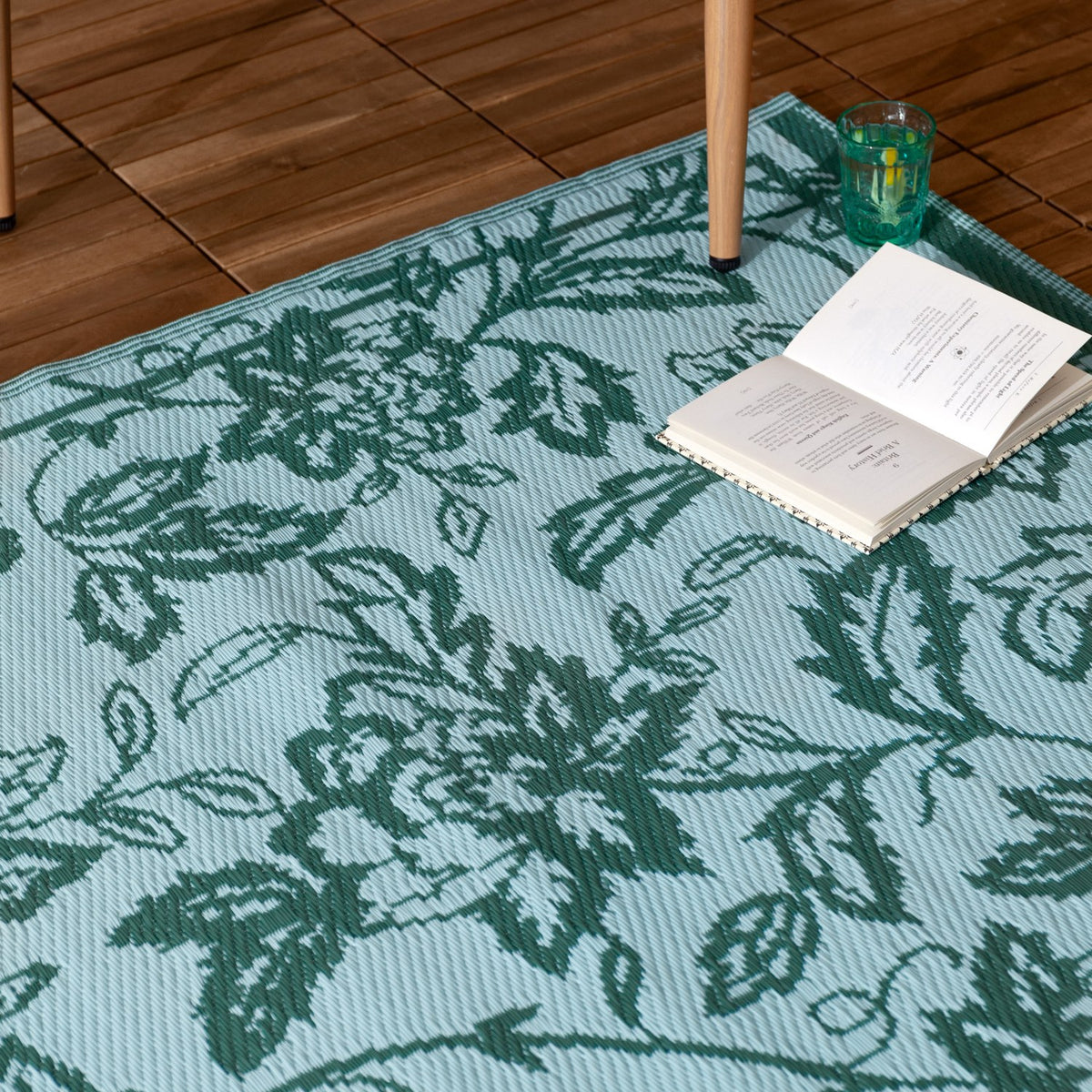 Anemone Jacquard Floral Rug in Teal/Mineral, an outdoor rug with a woven jacquard floral pattern in teal-green on a mineral-blue ground, shown on decking with a book and glass.
