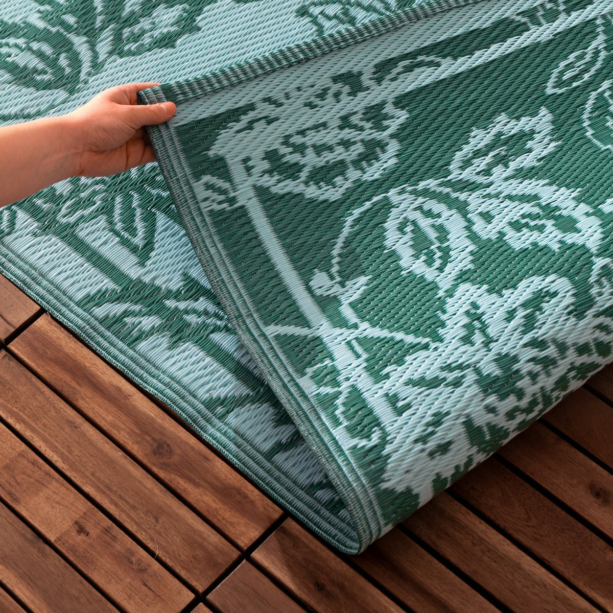 Hand lifting the corner of the Anemone Jacquard Floral Rug in teal and mineral-blue, showing the jacquard floral weave and bound edge on a wooden deck.