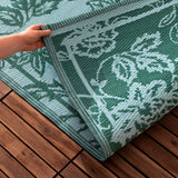 Hand lifting the corner of the Anemone Jacquard Floral Rug in teal and mineral-blue, showing the jacquard floral weave and bound edge on a wooden deck.