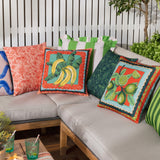 Anemone Reversible Polyester Cushion in coral-red with printed limes and scalloped teal, peach and gold border, shown among patterned cushions on an outdoor sofa.
