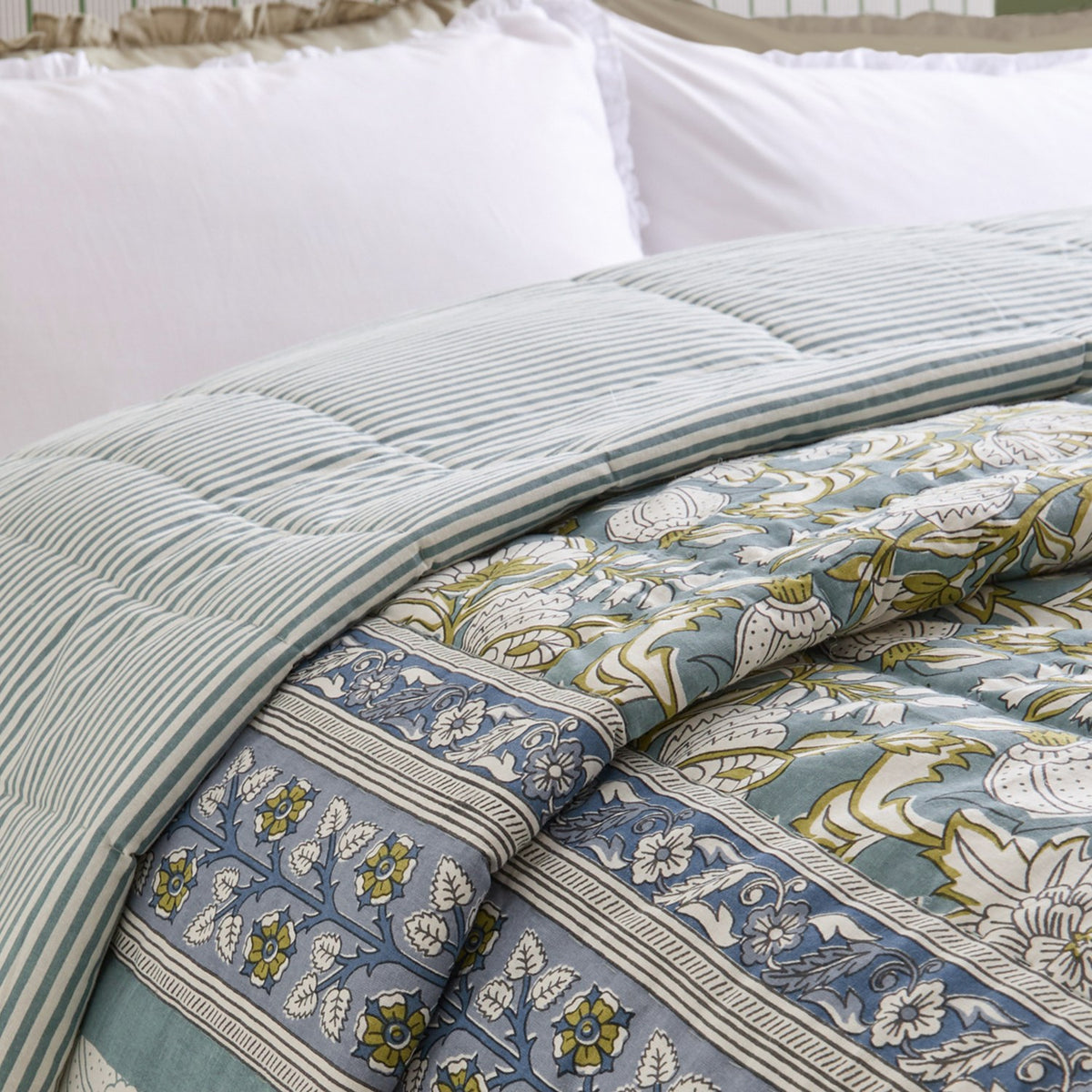 Annabelle reversible cotton bedspread in blue on a bed, showing the striped reverse folded back to reveal the floral, foliage-patterned face with striped border.