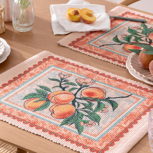 Anemone Floral Polyester Placemats in multicolour, rectangular woven mats with a peach branch print and orange floral border, shown styled on a wooden dining table with fresh peaches.