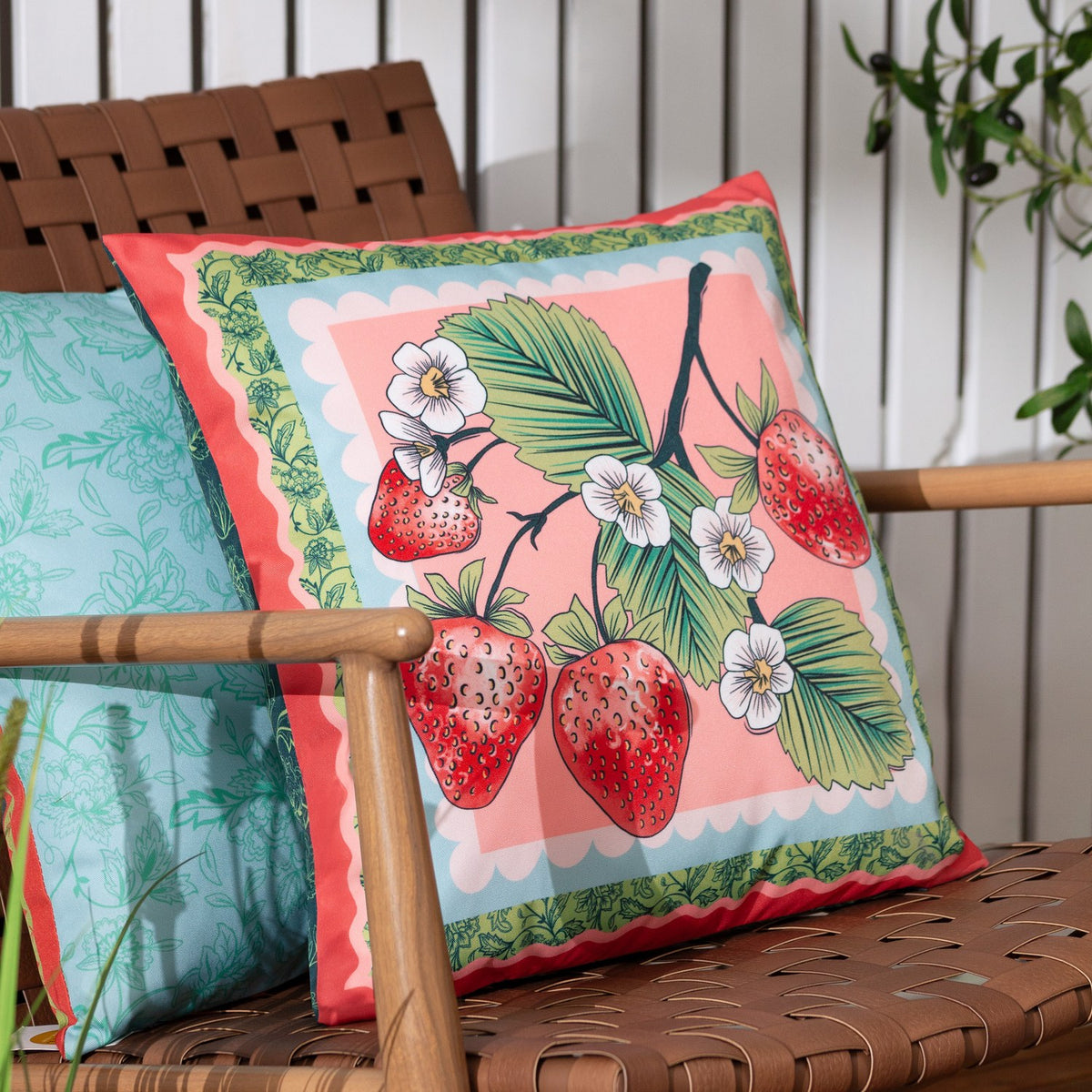 Anemone Reversible Polyester Cushion in Multi, coral-pink square cushion with strawberry, leaf and white flower print and scalloped green border, shown on an outdoor chair.
