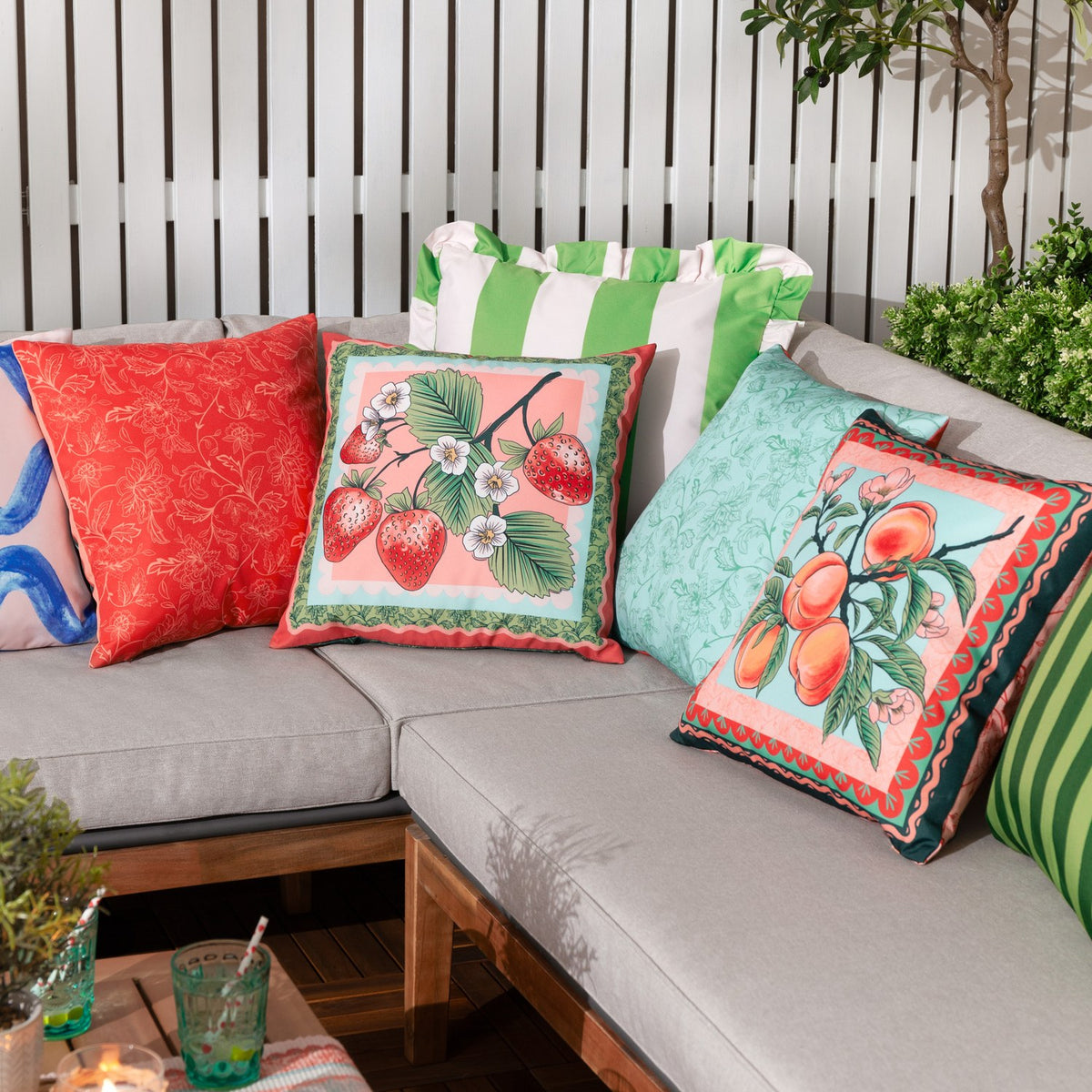 Anemone Reversible Polyester Cushion in Multi with a strawberry print on coral-pink, styled among patterned scatter cushions on an outdoor sofa.