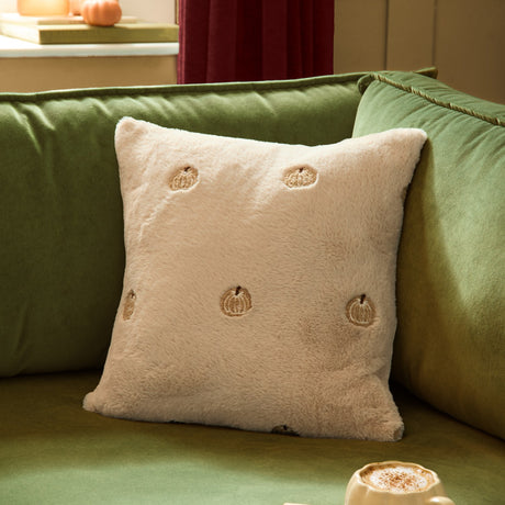Square faux fur cushion in natural cream with embroidered gold and tan pumpkin motifs, shown on a green sofa in a living room setting.