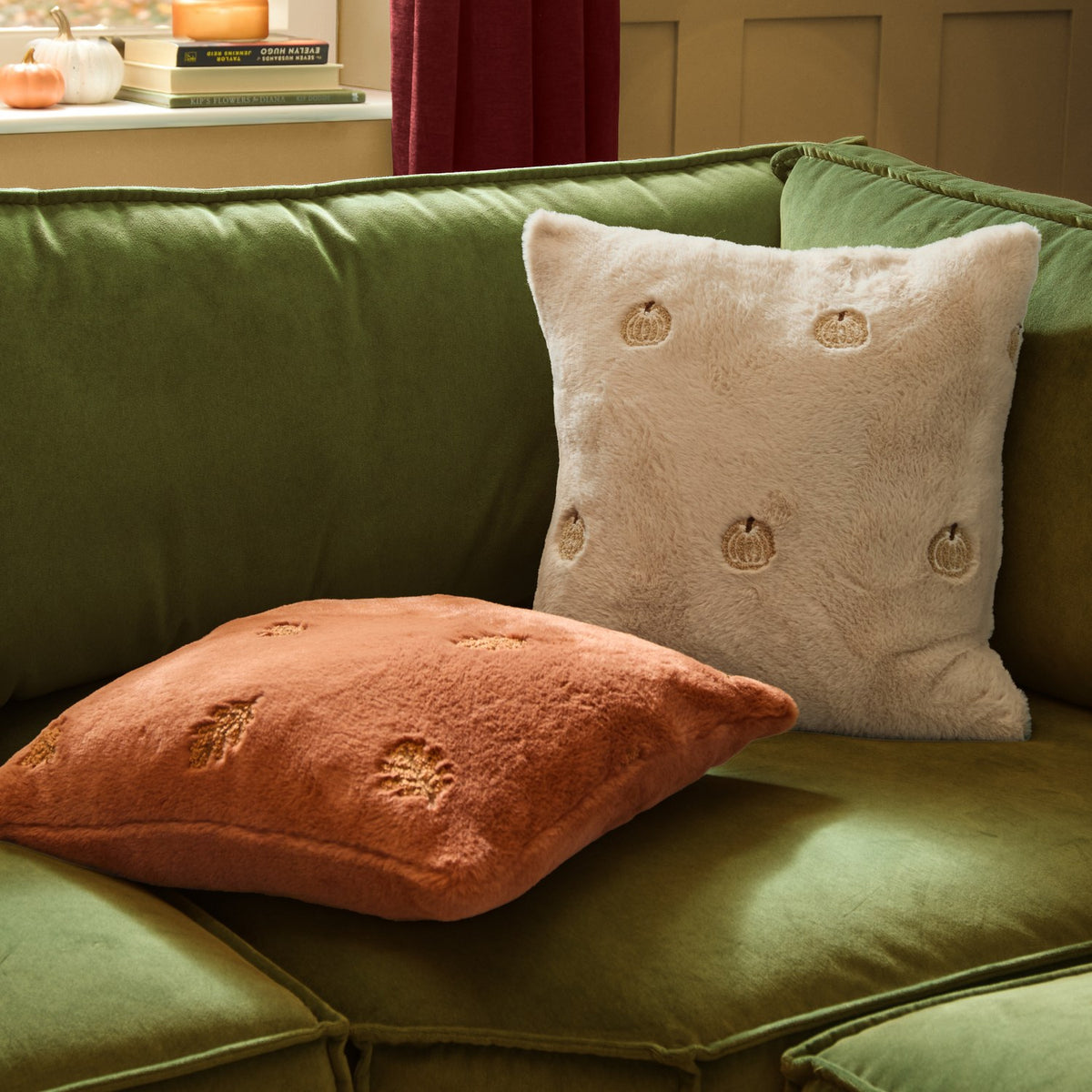 Square natural cream faux fur cushion with embroidered gold-tan pumpkin motifs, shown on a green velvet sofa beside a terracotta cushion.