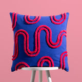 Archie Abstract Cotton Blend Cushion in cobalt blue with hand-tufted pink swirl and arch pattern, square 45 x 45cm design, shown on a stool against a pink background.