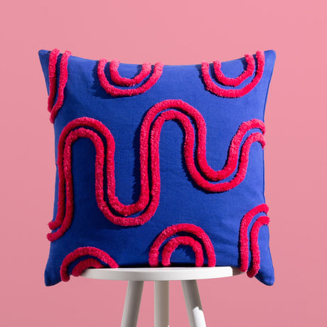Archie Abstract Cotton Blend Cushion in cobalt blue with hand-tufted pink swirl and arch pattern, square 45 x 45cm design, shown on a stool against a pink background.