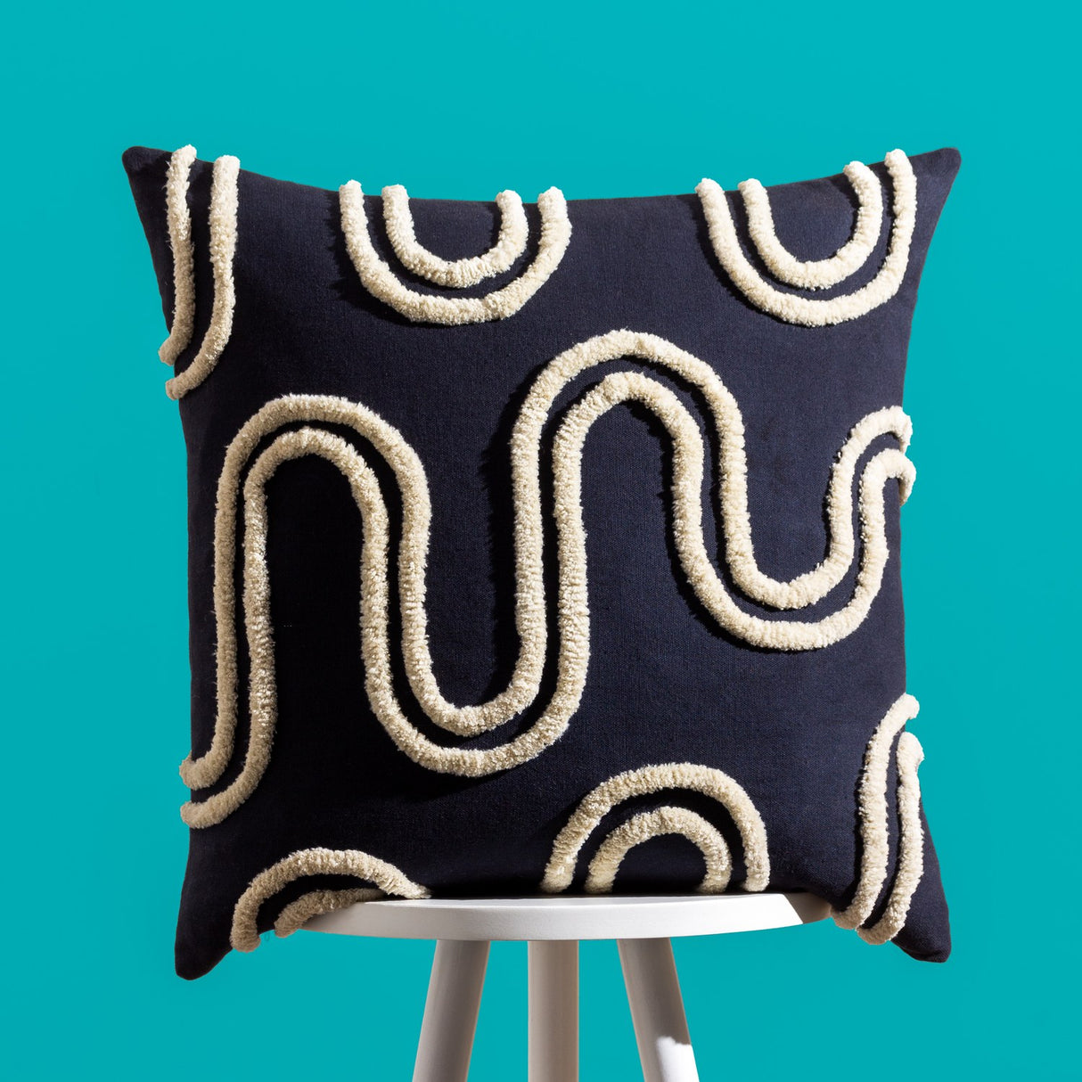 Archie Abstract Cotton Blend Cushion in Mono, a 45 x 45cm square cushion with tufted cream abstract swirl and arch patterns on a black cotton blend cover, shown on a stool against a teal background.