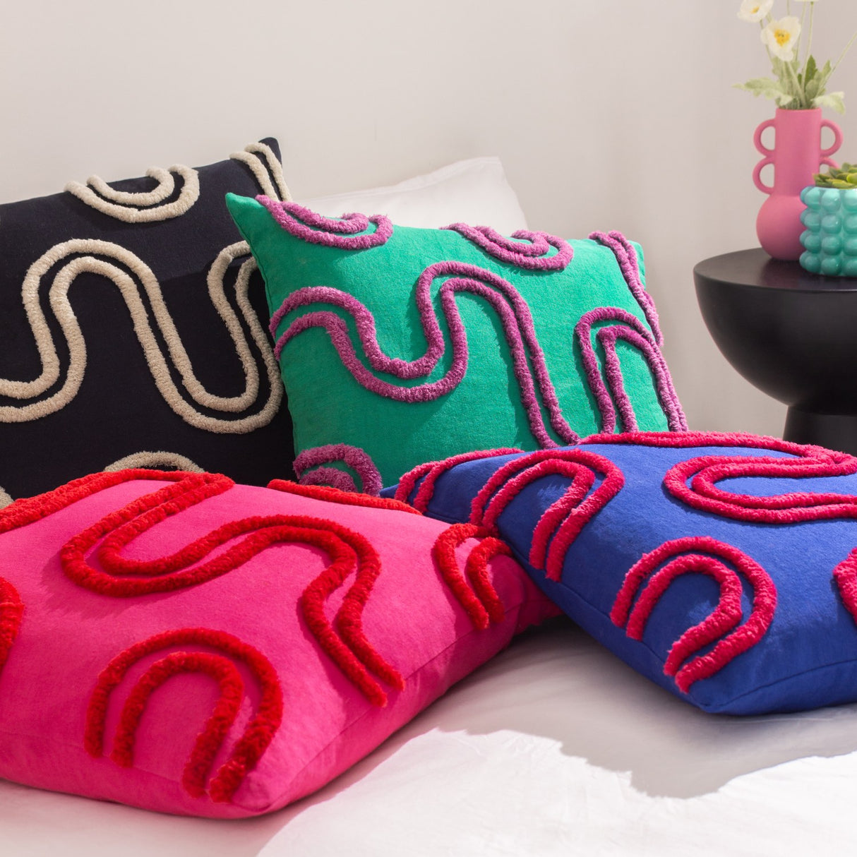 Archie Abstract Cotton Blend Cushion in Mono, black with cream tufted swirl design, shown with pink, green, and blue patterned cushions on a bed.