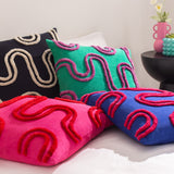 Archie Abstract Cotton Blend Cushion in Mono, black with cream tufted swirl design, shown with pink, green, and blue patterned cushions on a bed.
