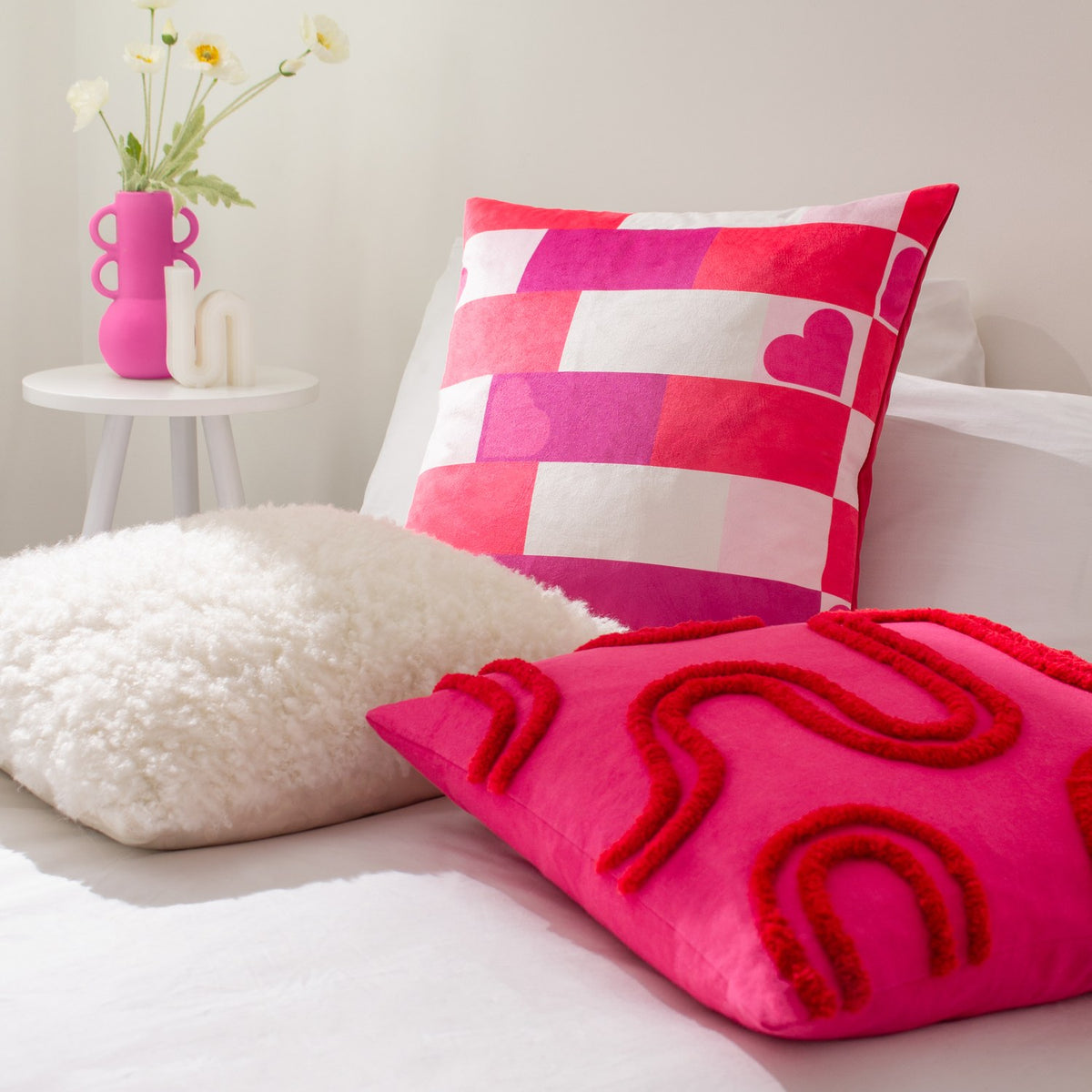 Archie Abstract Cotton Blend Cushion in pink with red tufted looping design, styled on a bed alongside a pink and white checked heart-print cushion and cream faux fur cushion.
