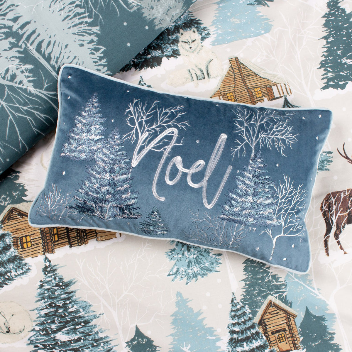 Rectangular blue velvet cushion from the Arcticus range, embroidered with frosted trees, bare branches and "Noel" script, shown on winter-themed bedding.