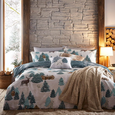 Arcticus Printed Animal Duvet Cover Set in blue and cream on a bedroom bed, featuring pine trees, log cabins, a stag and Arctic animals, styled with matching pillowcases and a faux-fur throw.