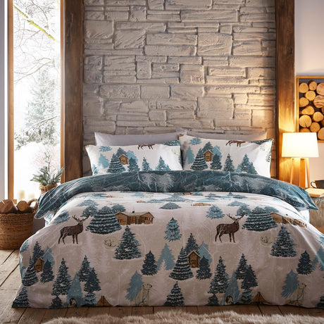 Arcticus Printed Animal Duvet Cover Set in blue and cream, showing stags, pine trees and log cabins on a bed against a stone-effect bedroom wall.