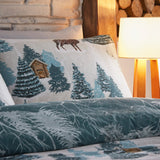 Arcticus Printed Animal Duvet Cover Set in blue and cream, showing a winter woodland scene with pine trees, a log cabin and a stag, styled on a bed beside a lit lamp.