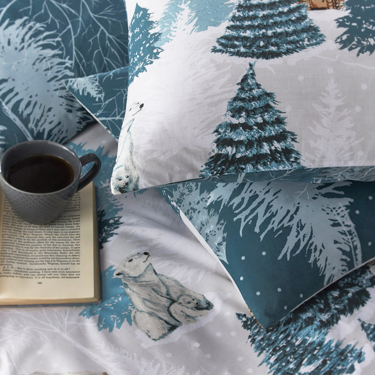 Arcticus Printed Animal Duvet Cover Set in blue and white, showing pillowcases with polar bears and snowy pine trees on a bed beside a mug and open book.