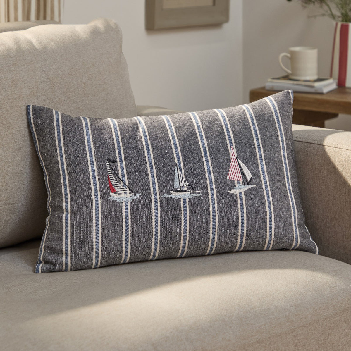Abersoch Regatta rectangular cushion in navy striped cotton with cream and blue lines, featuring three embroidered sailboats, shown on a beige sofa.