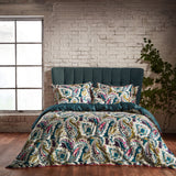 Aretha Cotton Floral Duvet Cover Set in teal and olive, styled on a bed with matching pillowcases in a paisley print of teal, plum, pink and ivory tones, against a whitewashed brick wall.