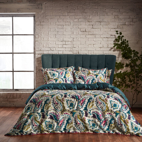 Aretha Cotton Floral Duvet Cover Set in teal and olive, styled on a bed with matching pillowcases in a paisley print of teal, plum, pink and ivory tones, against a whitewashed brick wall.