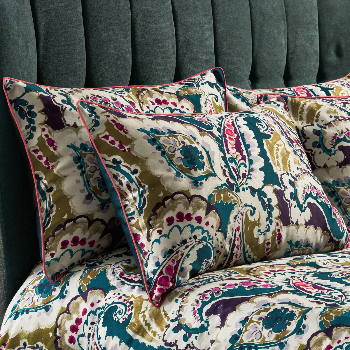Aretha Floral Cotton Pillowcase Pair in teal, olive, purple and magenta paisley print on a cream ground, with contrast pink piped edges, styled on a bed against a teal velvet headboard.