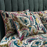 Aretha Floral Cotton Pillowcase Pair in teal, olive, purple and magenta paisley print on a cream ground, with contrast pink piped edges, styled on a bed against a teal velvet headboard.