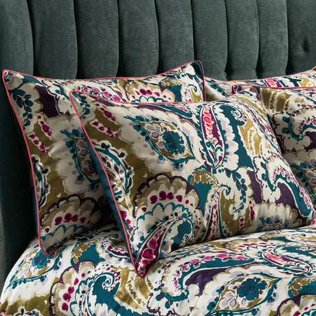 Aretha Floral Cotton Pillowcase Pair in teal, olive, purple and magenta paisley print on a cream ground, with contrast pink piped edges, styled on a bed against a teal velvet headboard.