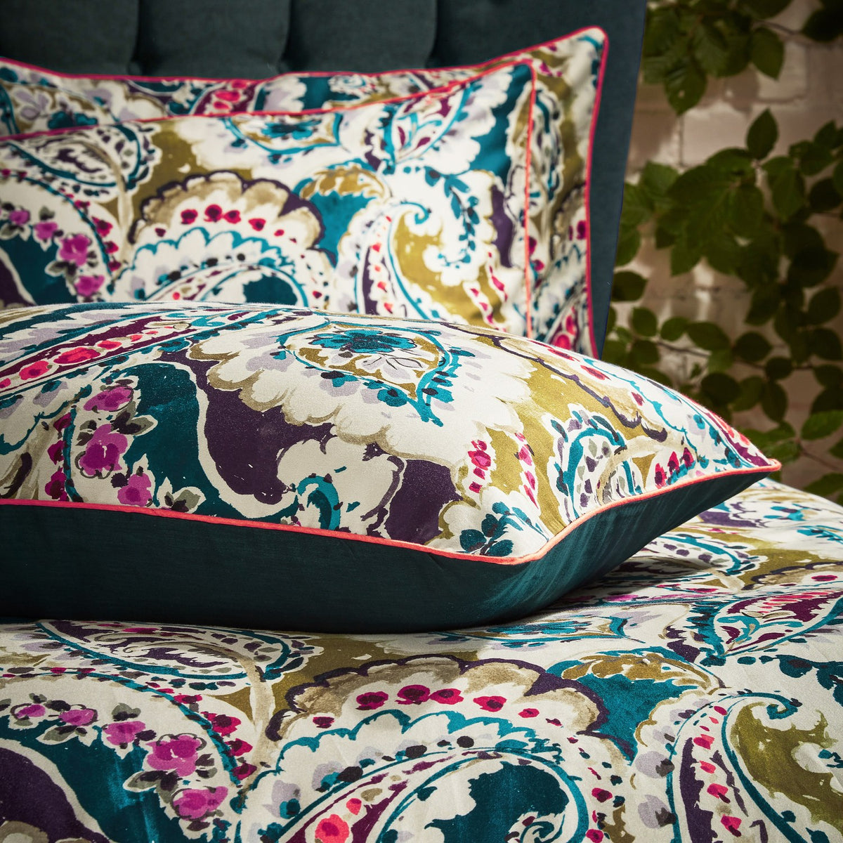 Aretha Floral Cotton Pillowcase Pair in teal/olive, showing large-scale paisley print in teal, olive, purple and magenta on cream cotton with coral piping, styled on a bed.