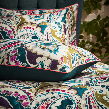 Aretha Floral Cotton Pillowcase Pair in teal/olive, showing large-scale paisley print in teal, olive, purple and magenta on cream cotton with coral piping, styled on a bed.