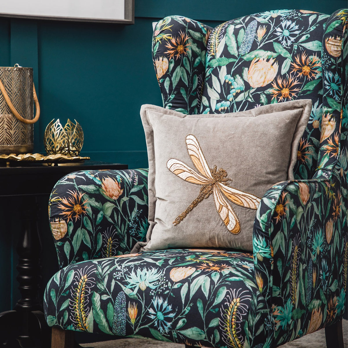 Aria Animal Velvet Cushion in grey with gold-tone embroidered dragonfly motif, shown on a floral-print wingback armchair in a dark teal room setting.
