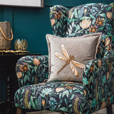 Aria Animal Velvet Cushion in grey with gold-tone embroidered dragonfly motif, shown on a floral-print wingback armchair in a dark teal room setting.