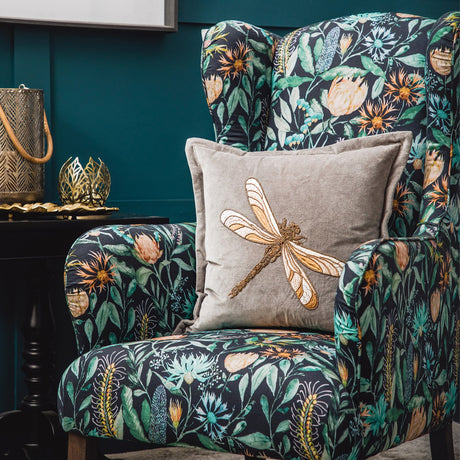 Aria Animal Velvet Cushion in grey with gold-tone embroidered dragonfly motif, shown on a floral-print wingback armchair in a dark teal room setting.