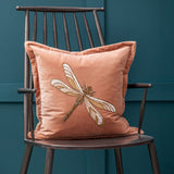 Aria Animal Velvet Cushion in dusky pink velvet with an embroidered gold and cream dragonfly design, shown propped on a dark metal spindle-back chair against a teal wall.
