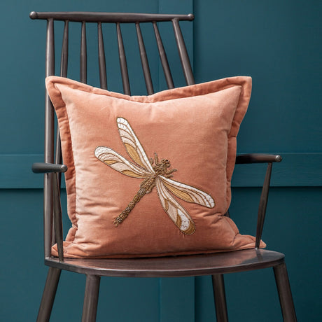 Aria Animal Velvet Cushion in dusky pink velvet with an embroidered gold and cream dragonfly design, shown propped on a dark metal spindle-back chair against a teal wall.
