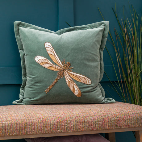 Aria Animal Velvet Cushion in teal, 50 x 50cm square with flanged edge, featuring a beaded dragonfly in amber and gold tones, shown on a bench against a dark teal panelled wall.