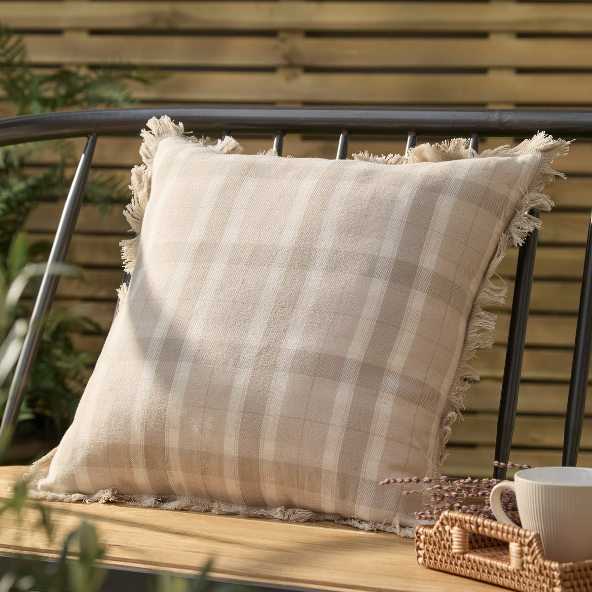 Arrochar Check Linen Cushion in Nut, a 50 x 50cm square cotton-linen cushion with oatmeal and off-white check and self-fringed edges, shown on a bench in an outdoor garden setting.