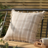 Arrochar Check Linen Cushion in Nut, a 50 x 50cm square cotton-linen cushion with oatmeal and off-white check and self-fringed edges, shown on a bench in an outdoor garden setting.