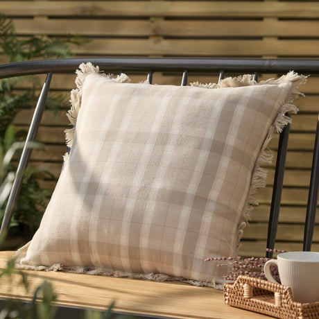 Arrochar Check Linen Cushion in Nut, a 50 x 50cm square cotton-linen cushion with oatmeal and off-white check and self-fringed edges, shown on a bench in an outdoor garden setting.