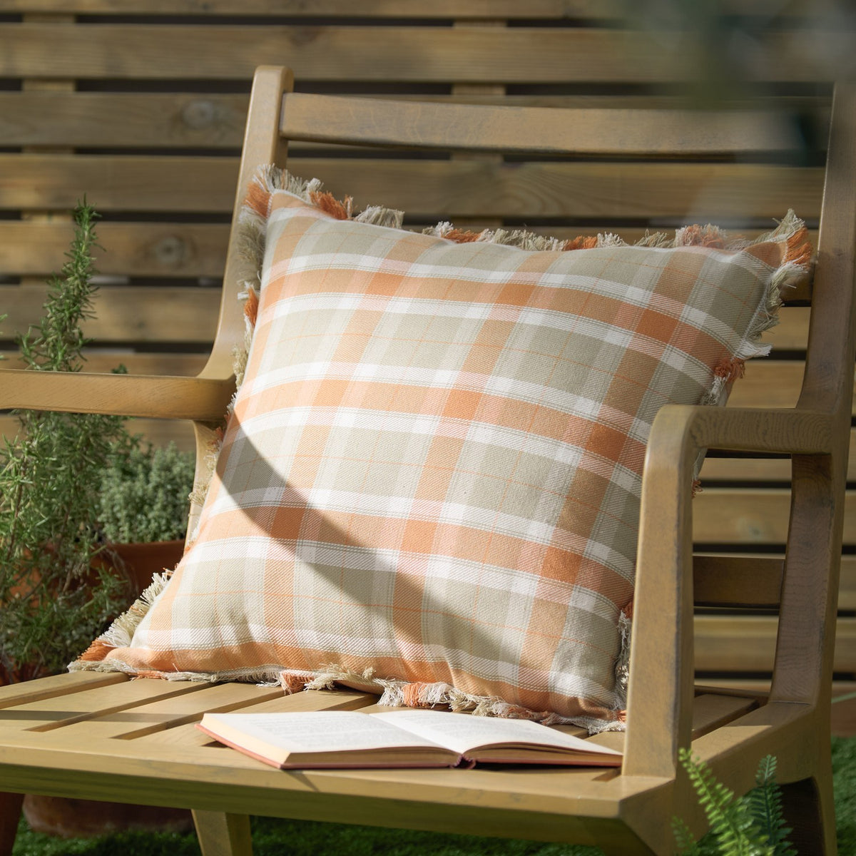 Arrochar Check Linen Cushion in Russet, a 50x50cm square cushion with an orange, taupe and off-white tartan check and self-fringed edging, shown on a wooden garden bench.