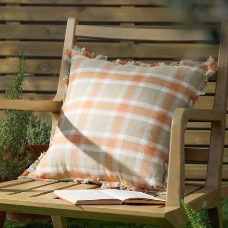 Arrochar Check Linen Cushion in Russet, a 50x50cm square cushion with an orange, taupe and off-white tartan check and self-fringed edging, shown on a wooden garden bench.