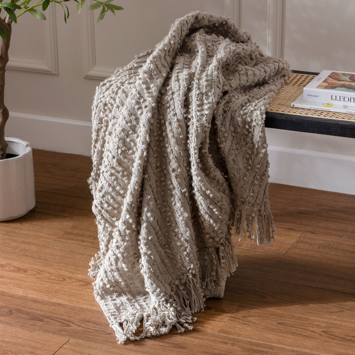 Arvo Plain Cotton Throw in feather grey with textured boucle stripes and fringed edges, draped over a bench beside a potted plant on a wooden floor.