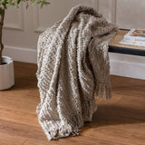 Arvo Plain Cotton Throw in feather grey with textured boucle stripes and fringed edges, draped over a bench beside a potted plant on a wooden floor.