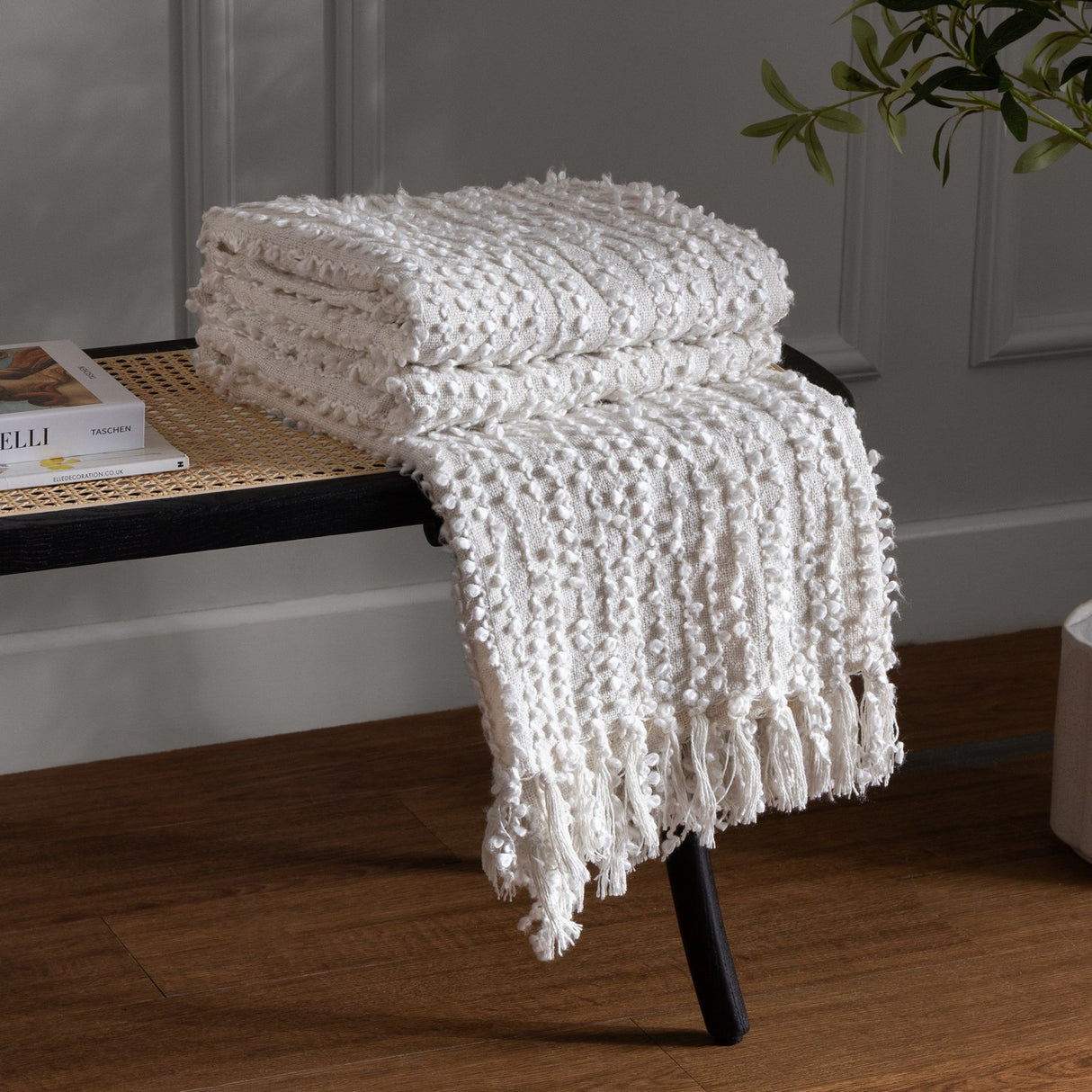 Arvo Plain Cotton Throw in natural cream, folded with textured boucle rows and fringed edge, draped over a black rattan bench beside books and a plant.