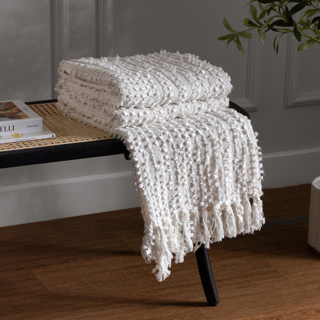 Arvo Plain Cotton Throw in natural cream, folded with textured boucle rows and fringed edge, draped over a black rattan bench beside books and a plant.