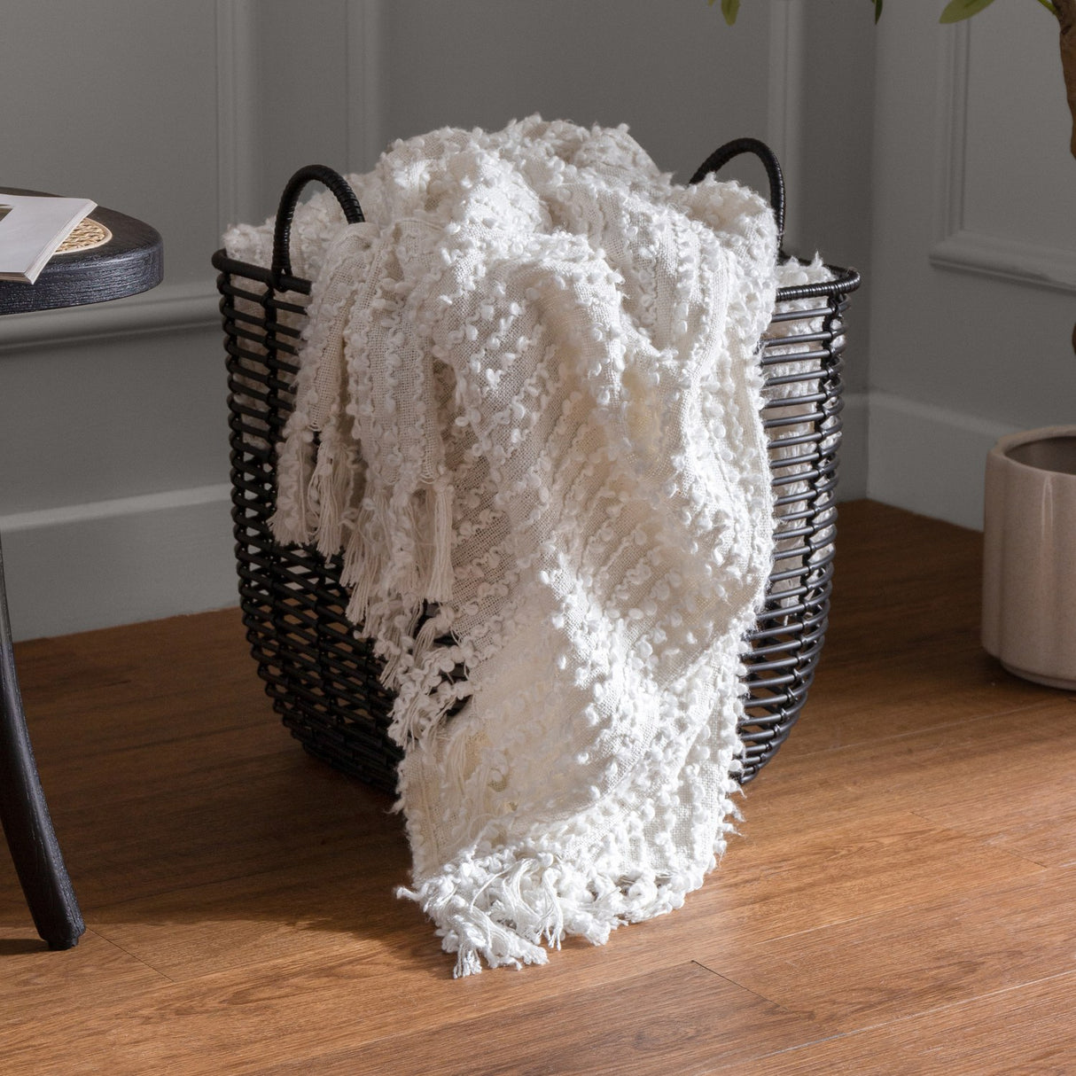 Arvo Plain Cotton Throw in natural white, woven with textured boucle rows, draped over a black wire storage basket with tasselled fringe trailing onto a wooden floor.