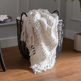 Arvo Plain Cotton Throw in natural white, woven with textured boucle rows, draped over a black wire storage basket with tasselled fringe trailing onto a wooden floor.