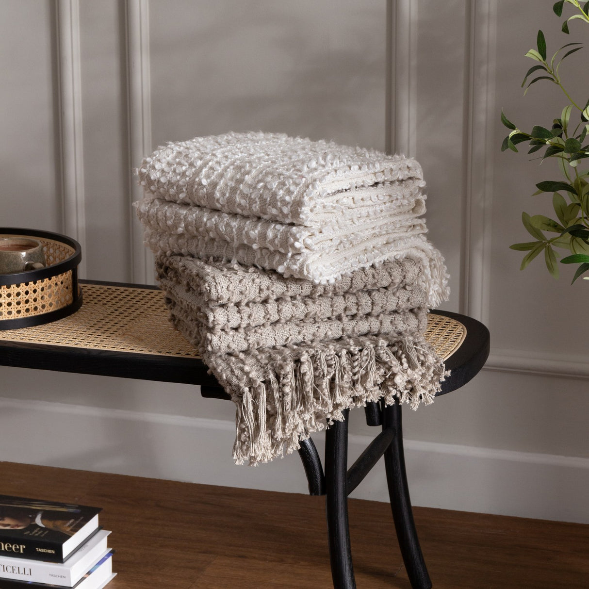 Arvo Plain Cotton Throw in natural, folded with textured boucle rows and fringed edge, stacked on a black cane bench beside books and a plant.