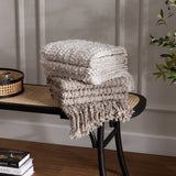 Arvo Plain Cotton Throw in natural, folded with textured boucle rows and fringed edge, stacked on a black cane bench beside books and a plant.